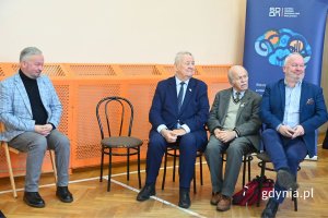 Czterej mężczyźni siedzący na krzesłach na tle beżowo-pomarańczowej ściany