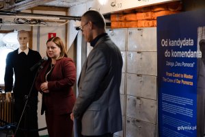 Kobieta w wiśniowej marynarce przy mikrofonie, obok manekin