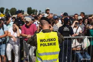 Żołnierz odwr&oacute;cony plecami w kamizelce z napisem Wojsko Polskie na tle gromady ludzi