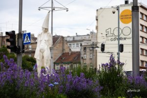 Rzeźba przedstawiająca kobietę z tr&oacute;jkątną czapką na głowie, stoi pomiędzy zielenią, w tle budynek z muralem "100 Gdynia. Zaczeło się od morza"
