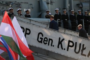 Ministrowie obrony państw Grupy Wyszehradzkiej wchodzą na pokład okrętu ORP "Gen. K. Pułaski". Na pierwszym planie powiewają flagi Polski, Węgier, Słowacji i Czech. Na okręcie w szeregu i na baczność stoją żołnierze Marynarki Wojennej // fot. Michał Puszczewicz