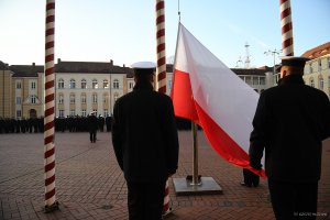 Dwaj mężczyźni w mundurach marynarskich stoją tyłem do fotografa, na maszt wciągana jest flaga Polski.