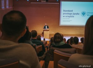 widoczne osoby na sali wykładowej, trwający wykład, osoba przemawia na tle prezentacji