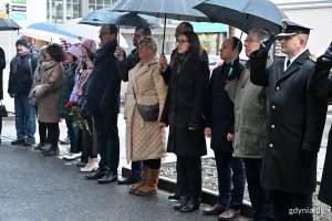 Grupa osób stoi pod parasolami na chodniku w szeregu.