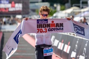 Zawodnik trzymający wstęgę z napisem Enea IRONMAN 70.3 Gdynia, w tle widoczna infrastruktura mety/startu. 