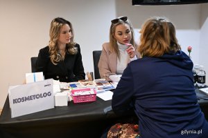 Trzy kobiety przy stoliku z tabliczką "kosmetolog", jedna z kobiet odwrócona plecami, pomieszczenie