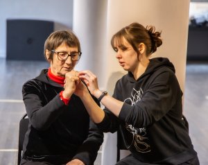 Dwie siedzące kobiety, jedna pomaga drugiej ułożyć dłońmi gest