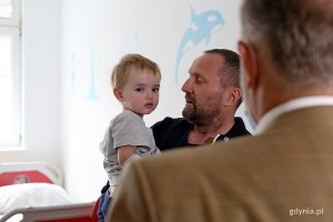 Prezydent Gdyni widziany od tyłu w pokoju szpitalnym. na przeciw stoi tata i trzyma na ręku małego chłopca.