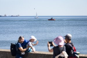 Po morzu płynie motorówka i jacht, na pierwszym planie ludzie się fotografują. 