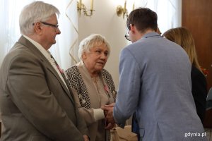 Mężczyna w garniturze ściska dłoń starszej pani, obok inny mężczyzna
