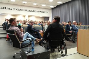 Kobieta na w&oacute;zku, mężczyzna z mikrofonem na pierwszym planie, w tle studenci na sali 