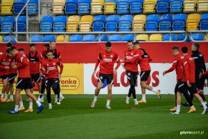 Zawodnicy w czerwonych bluzach i czarnych spodenkach podczas treningu na murawie stadionu. W tle żółto-niebieskie trybuny. 