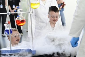 Dwoje młodych chłopców w białych fartuchach przy stanowisku do eksperymentów. Obok nich znajdują się próbówki.  