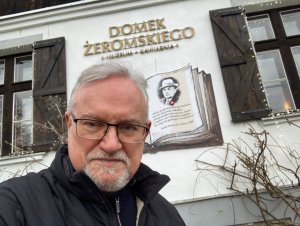 Mężczyzna na tle białej ściany z oknami w brązowych okiennicach ozdobionej grafiką książki z twarzą Stefana Żeromskiego i szyldem z nazwą muzeum