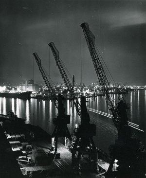 Fotografia przedstawia port w Gdyni po zmroku. Na pierwszym planie widok z góry na nabrzeże Fińskie z trzema, ustawionymi w rzędzie dźwigami portowymi. Na dalszym planie widoczny jest oświetlony port wewnętrzny, z lewej początek basenu im. Marsz. Józefa Piłsudskiego, w którym zacumowany jest statek handlowy. Widoczny przy nabrzeżu Indyjskim przed elewatorem zbożowym. Po prawej stronie, widoczne dźwigi portowe i światła portu w Gdynia. Fotografia pochodzi z archiwum cyfrowego Muzeum Miasta Gdyni // fot. Halina Wasielke