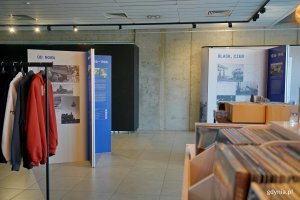 Ekspozytory z wystawą &bdquo;Gdynia x 10&rdquo; w Galerii Gdyńskiego Centrum Filmowego im. Leszka Kopcia stojące obok wieszak&oacute;w z bluzami i p&oacute;łek z płytami