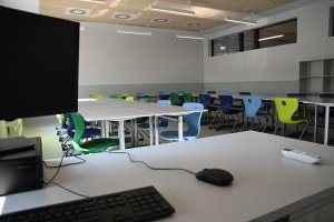 Na pierwszym planie komputer na biurku nauczyciela w tle ławki ustawione w kształt litery U z kolorowymi krzesłami