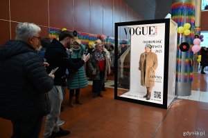 Korytarz Polsat Plus Areny Gdynia, dekoracja do fotografii z napisem "Vogue", wewnątrz pozuje seniorka, kilkanaście osób obok
