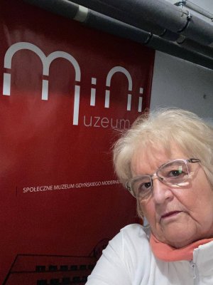 Kobieta na tle czerwonego plakatu z nazwą muzeum wiszącego na białej ścianie