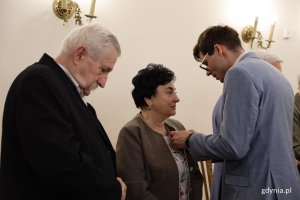 Mężczyzna w garniuturze przypina medal starszej pani w beżowej marynarce