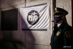 Tablica pamiątkowa na fasadzie budynku Urzędu Miasta przed odsłonięciem - osłonięta białą flagą z czarnym znakiem Grudnia 1970. Przy tablicy stoi strażniczka miejska w mundurze // fot. Karol Stańczak