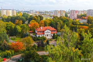 Widok z lotu ptaka na budynek z pomarańczowym dachem, który jest otoczony drzewami i krzewami w jesiennej szacie. W tle pobliskie bloki // fot. Michał Piszczewicz