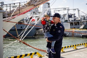 Marynarz z chłopcem na rękach stoi na nabrzeżu obok żaglowca.