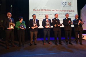 Grupa osób stojących na scenie i mających za plecami biały ekran