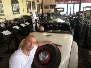 Mężczyzna przy samochodzie w kolorze kremowym, w tle widoczne motocykle i muzealne tabliczki