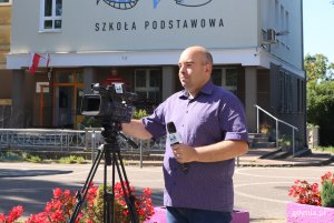 Upalny dzień, mężczyzna przygotowuje kamerę video do nagrania, w tle budynek Zespolu Szkół Ogólnokształcących nr 6 w Gdyni z grafiti na fasadzie