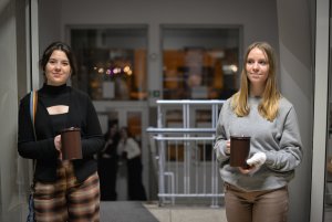 Dwie dziewczyny stojące po bokach drzwi, w tle klatka schodowa i drzwi wejściowe do szkoły