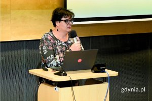 Kobieta stojąca za pulpitem z laptopem i mówiąca do mikrofony