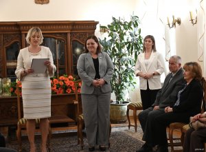 Kobieta w biały stroju, obok kobieta w szarej marynarce, w tle inne osoby