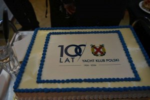 Biały tort z niebieskimi zdobieniami i napisem „100 lat Yacht Klub Polska 1924-2024” 