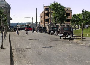 Ulica z zaparkowanymi samochodami, na której stoi kilka osób, w tle jedzie autobus i stoi budynek.