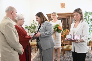 Kobieta w szarej marynarce ściska dłoń kobiety w czerwonej sukience, obok inne osoby