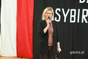 Kobieta stojąca i mówiąca do mikrofonu na tle granatowej dekoracji z motywem polskiej flagi