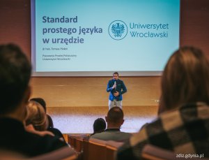 widoczne osoby na sali wykładowej, trwający wykład, osoba przemawia na tle prezentacji
