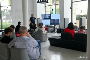 Na pierwszym planie uczestnicy spotkania w Muzeum miasta Gdyni siedzący na sofach. Z tyłu: ornitolog Jakub Typiak wygłaszający prelekcję