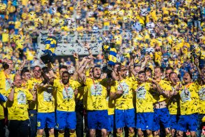 Piłkarze Arki Gdynia w żółto-niebieskich sportowych strojach na tle spadającego konfetti. Jeden z zawodników trzyma puchar. 