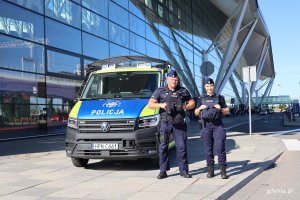 Dwie osoby z policji na tle samochodu służbowego w niebiesko-żółtym w tle budynek lotniska
