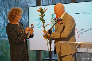 Wojciech Bonisławski w eleganckim garniturze wręczający Nagrodę Marszałka Województwa Pomorskiego i kwiata Caren Garfen