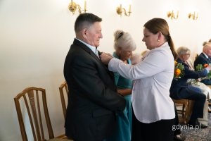 Kobieta w różowej marynarce przypina medal starszemu mężczyźnie