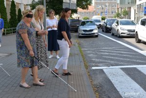 Kobieta z zasłoniętymi oczami i z baiłą laską stoi przed przejściem dla pieszych.