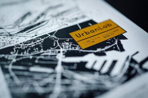 Grafika prezentuje żółte logo UrbanLab na tle biało-czarnej mapy Gdyni