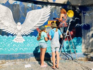 Dwie osoby stoją przy muralu tyłem do obiektywu, jedna osoba z pędzlem stoi na drabinie i kończy malowanie muralu