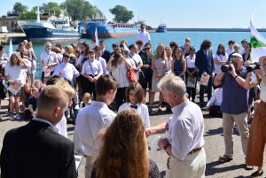 Grupa uczniów w tle wody zatoki i zacumowane statki oraz wejście do portu