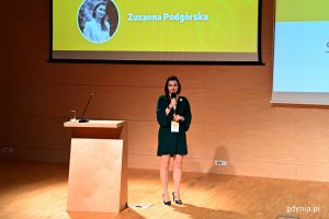 Zuzanna Podgórska, popularyzatorka nauki i prezes Stowarzyszenia Rzecznicy Nauki podczas Gdynia Explory Week. Stoi obok mównicy. W tle telebim ze zdjęciem prelegentki // fot. Magdalena Czernek