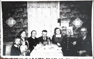 Fotografia &ndash; sądząc po stylu ubioru, fryzurach i charakterystycznym wystroju wnętrza mogła zostać wykonana w latach 40. lub 50. XX wieku. W czarno-białym kadrze ujęto kilka os&oacute;b w r&oacute;żnym wieku, w tym dziecko z lalką i mężczyznę w mundurze, siedzących przy wsp&oacute;lnym stole &ndash; zapewne rodzinę uchwyconą podczas uroczystego spotkania.