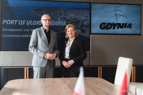 Sala konferencyjna w Urzędzie Miasta Gdyni, wiceprezydent Austen pozuje do zdjęcia z ambasador Mimozą Halimi. Za nimi dwa wyświetlacze ze zdjęciami portu we Wlorze oraz logotypem Gdyni.